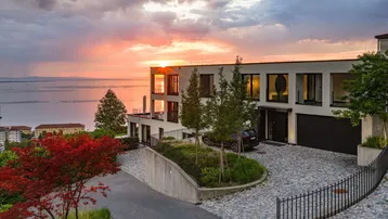 Moderne Villa mit Seeblick bei Sonnenuntergang, umgeben von grüner Landschaft und gepflasterter Auffahrt.
