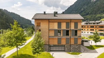 Modernes Gebäude mit Holzfassade und Schiebeelementen vor den Fenstern, umgeben von grüner Landschaft und Bergen.