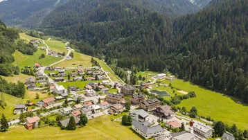 Luftaufnahme eines Dorfes in einem grünen Tal, umgeben von bewaldeten Bergen und Sportanlagen.