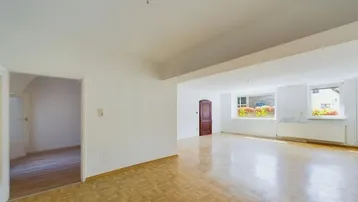 Helles, leeres Zimmer mit Parkettboden und Fenstern, die den Blick nach draußen freigeben.