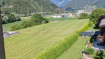 Weitläufiger Blick über grüne Felder und Hecken auf die umliegende Berglandschaft unter blauem Himmel.