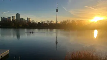 Sonnenuntergang über einem See mit Stadtpanorama und Ruderbooten im Vordergrund.