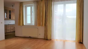 Helles Wohnzimmer mit Laminatboden, großen Fenstern und Blick in die angrenzende Küche.