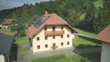 Haus mit Ziegeldach, Photovoltaikanlage und Holzbalkon, umgeben von grüner Landschaft.