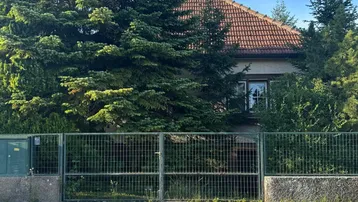 Hausfassade hinter einem Zaun und hohen Bäumen, mit einem roten Ziegeldach und grüner Umgebung.