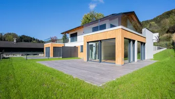Moderne Villa mit Holzfassade, großer Holzterrasse und gepflegtem Rasen unter blauem Himmel.