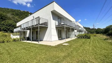 Modernes Wohngebäude mit mehreren Balkonen und einer gepflegten Rasenfläche vor einer grünen Hügellandschaft.
