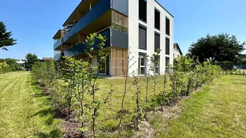 Modernes Mehrfamilienhaus mit Balkonen und gepflegter Grünanlage unter blauem Himmel.