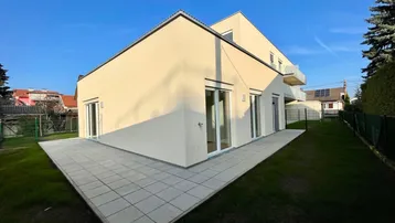 Modernes Gebäude mit großer gepflasterter Terrasse und angrenzendem Rasenbereich unter blauem Himmel.