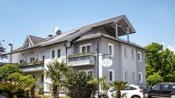 Modernes Gebäude mit mehreren Balkonen, Palmen im Vorgarten und einem Hinweisschild.