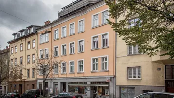 Mehrstöckiges Gebäude mit Geschäften im Erdgeschoss und Wohnungen darüber unter bewölktem Himmel.