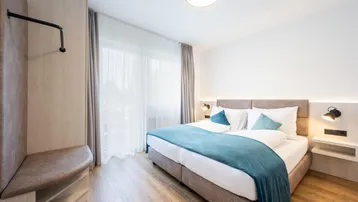 Modernes Schlafzimmer mit Doppelbett, Holzboden und großen Fenstern, die viel Tageslicht hereinlassen.