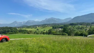 Panoramablick über grüne Felder und Wälder mit Bergen im Hintergrund und einem roten Auto.