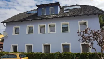 Ansicht des Hauses mit hellblauer Fassade, dunklem Dach und gepflegter Hecke im Vorgarten.