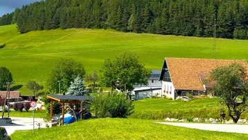 Ländliche Ansicht eines Hauses mit rotem Dach, umgeben von grünen Wiesen und bewaldeten Hügeln.