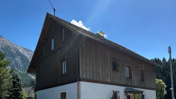 Traditionelles Haus mit Holzfassade und weißem Putz, umgeben von Bergen und grüner Natur, bei sonnigem Wetter.