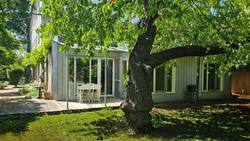 Modernes Haus mit Holzterrasse und Garten, umgeben von üppigem Grün und einem Obstbaum.