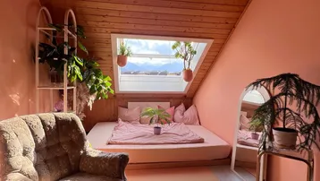 Gemütliches Schlafzimmer im Dachgeschoss mit Holzbalkendecke, rosa Wänden und einem großen Dachfenster mit Bergblick.