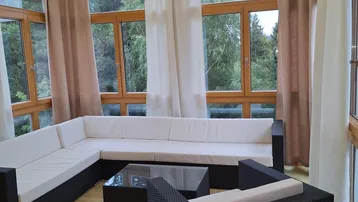 Heller Wintergarten mit großen Fenstern, Holzbalken und gemütlicher Sitzgruppe mit Blick ins Grüne.
