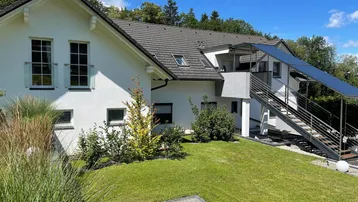 Weißes Einfamilienhaus mit Satteldach, gepflegtem Garten und Außentreppe unter blauem Himmel.