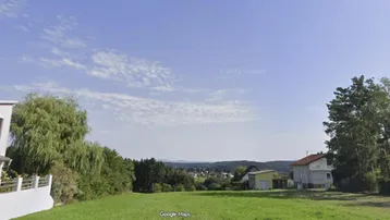 Weitläufige grüne Wiese mit Blick auf die umliegende Landschaft und entfernte Häuser unter blauem Himmel.