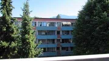 Blick von einem Balkon auf ein gegenüberliegendes Gebäude mit blauen Balkonen und Bergpanorama.