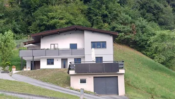 Modernes Haus am Hang mit Balkonen und integrierter Garage, umgeben von grünen Bäumen.