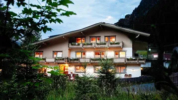 Mehrstöckiges Gebäude im alpinen Stil bei Dämmerung, umgeben von Bäumen und Natur.