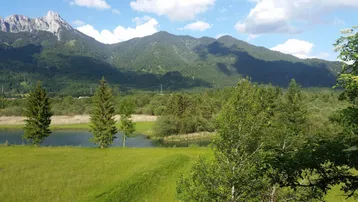 Panoramablick auf majestätische Berge, einen ruhigen See und üppige grüne Landschaft unter blauem Himmel.