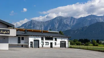 Modernes Geschäftsgebäude mit großem Parkplatz und beeindruckendem Alpenpanorama im Hintergrund unter blauem Himmel.