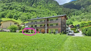 Mehrfamilienhaus in ländlicher Umgebung mit grünen Wiesen und bewaldeten Bergen im Hintergrund.