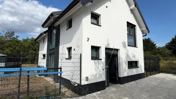 Modernes Einfamilienhaus mit weißer Fassade, dunklem Dach und gepflasterter Auffahrt unter blauem Himmel.