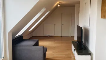 Helles Wohnzimmer im Dachgeschoss mit Holzboden, gemütlichem Sofa und Fernseher.