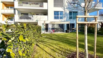 Modernes Apartmentgebäude mit gepflegtem Rasen, Hecken und einer Terrasse im Erdgeschoss.