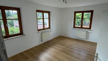 Geräumiges Zimmer mit hellem Laminatboden und zwei Fenstern mit Blick ins Grüne.