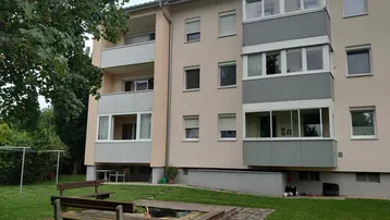 Außenansicht eines Mehrfamilienhauses mit Balkonen und einem gemeinschaftlichen Gartenbereich mit Sitzgelegenheiten.