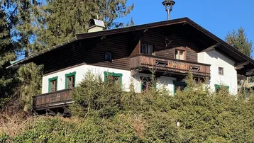Traditionelles alpenländisches Haus mit Holzbalkonen und weißer Fassade, umgeben von grünen Bäumen.