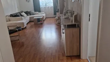 Helles Wohnzimmer mit Laminatboden, beigem Sofa und einem Fenster, das Tageslicht hereinlässt.