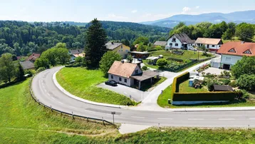 Luftaufnahme einer ländlichen Siedlung mit Häusern, Straßen und grünen Hügeln unter blauem Himmel.