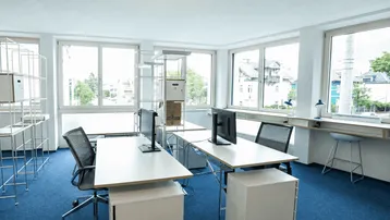 Helles und modernes Büro mit blauem Teppichboden, großen Fenstern und Blick ins Grüne.