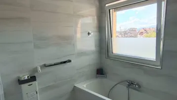 Modernes Badezimmer mit Badewanne, Toilette und Fenster, hell gefliest in Grautönen.