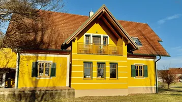 Gelbes Haus mit rotem Ziegeldach und einem Balkon im Obergeschoss. Grüne Fensterläden ergänzen die Fassade.