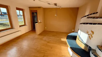 Heller Wohnbereich mit Holzboden und einem traditionellen Kachelofen. Große Fenster bieten viel Tageslicht.