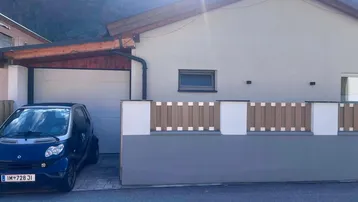 Modernes Haus mit Carport und Zaun, umgeben von einer beeindruckenden Berglandschaft.