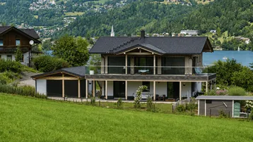 Modernes Haus mit dunklem Dach, Holzfassade und großem Balkon, eingebettet in eine grüne Hügellandschaft mit Seeblick.