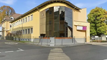 Gelbes Gebäude mit markanter, geschwungener Glasfassade an einer Straßenecke unter blauem Himmel.