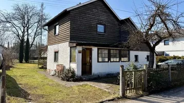 Vorderansicht eines Einfamilienhauses mit weißem Putz, dunkler Holzverkleidung und kleinem Vorgarten.