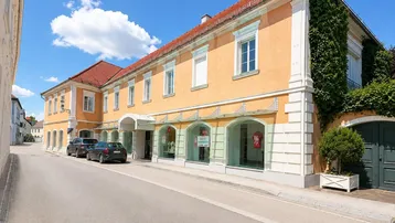 Historisches Gebäude mit ockerfarbener Fassade, roten Ziegeldächern und großen Schaufenstern im Erdgeschoss an einer Straße.