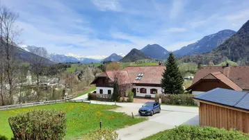 Idyllisches Haus mit Bergblick, umgeben von grünen Wiesen und Bäumen unter blauem Himmel.