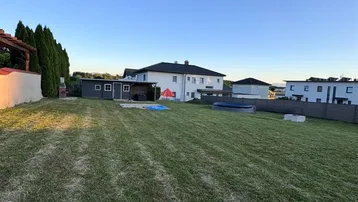 Großer Rasengarten mit Gartenhaus und einem aufblasbaren Pool im Hintergrund unter blauem Himmel.